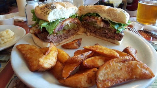 A plate with a halved burger topped with lettuce, tomato, and pickles, alongside crispy potato wedges.