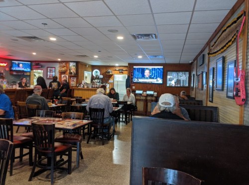 A casual restaurant interior with tables, patrons, and TVs showing news, featuring a warm, inviting atmosphere.