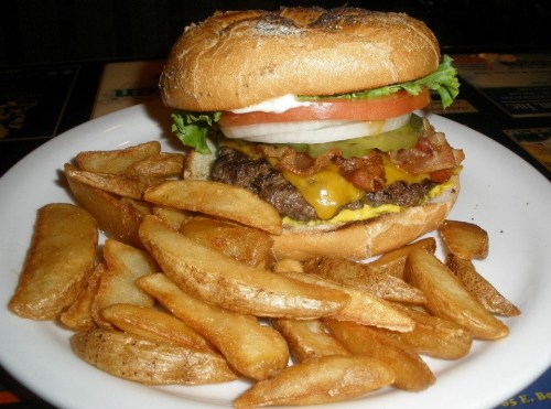A juicy cheeseburger with lettuce, tomato, pickles, and bacon, served with crispy potato wedges on a plate.
