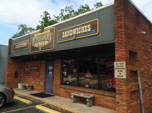 Exterior of a cafe with a brick facade, signs for "Sandwiches" and "Ruth's Cashews," and a parking sign nearby.