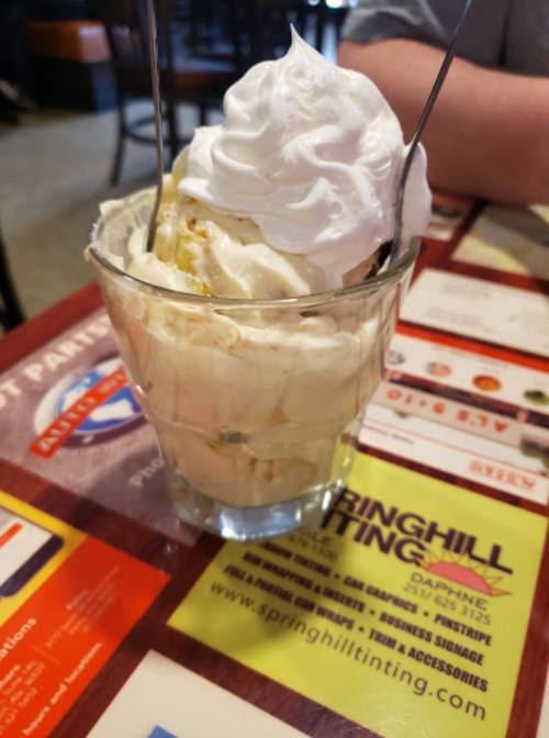 A glass dessert cup filled with creamy ice cream topped with whipped cream and two spoons.