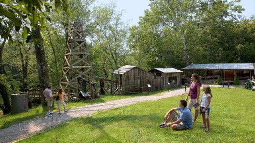 A group of people explores a historical site with wooden structures and a tall tower surrounded by trees.