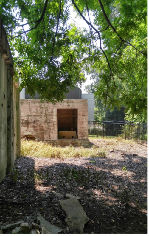 A weathered concrete structure surrounded by overgrown grass and trees, with a fence in the background.