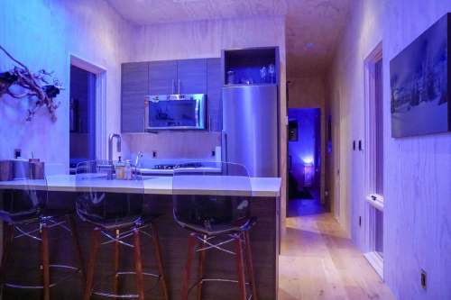 Modern kitchen with blue lighting, sleek cabinetry, and bar stools, leading to a hallway with ambient lighting.