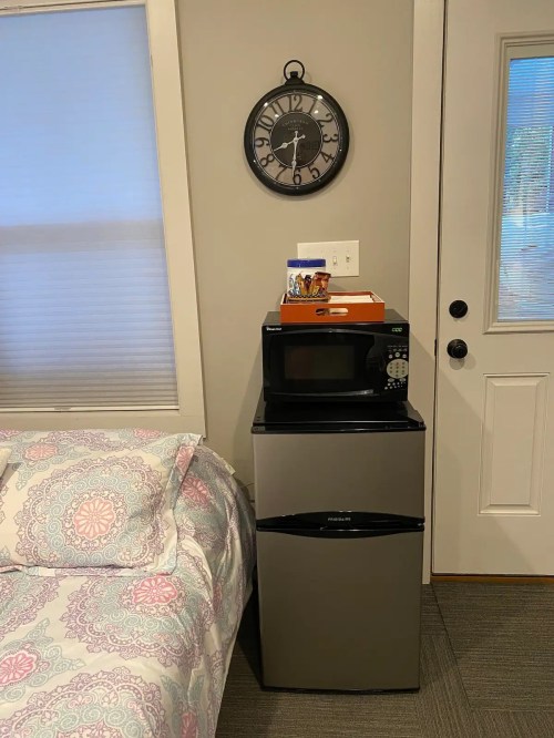 A small bedroom corner with a bed, a clock on the wall, and a microwave atop a mini fridge near a door.