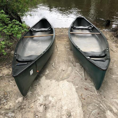 Two canoes sit on a sandy riverbank, facing each other, with trees and water in the background.