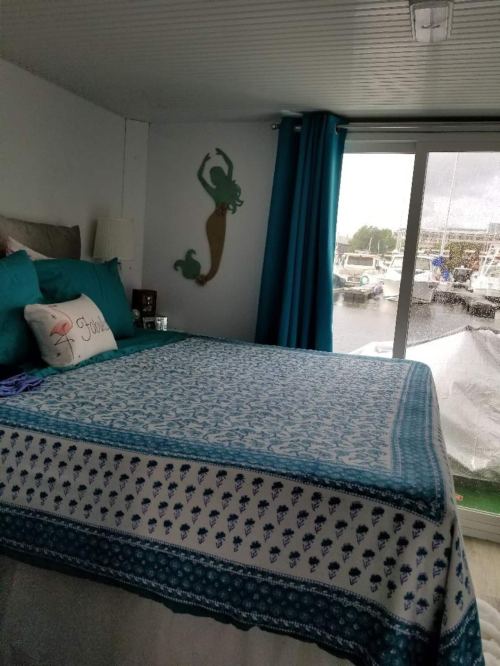 Cozy bedroom with a blue and white patterned bedspread, teal curtains, and a mermaid wall decoration overlooking a marina.