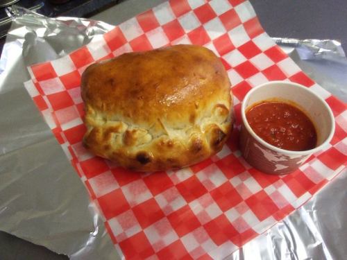 A golden-brown baked pastry on a red and white checkered paper, accompanied by a small cup of marinara sauce.