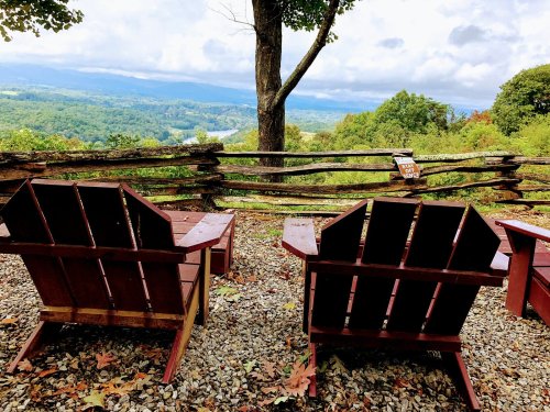 Two wooden chairs facing a scenic view of mountains and trees, with a rustic fence in the foreground.