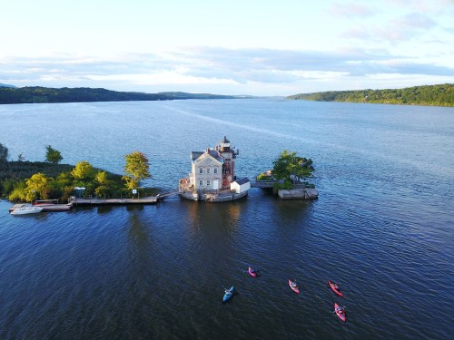 Aerial view of a small house on an island in a river, with kayakers paddling nearby under a clear sky.