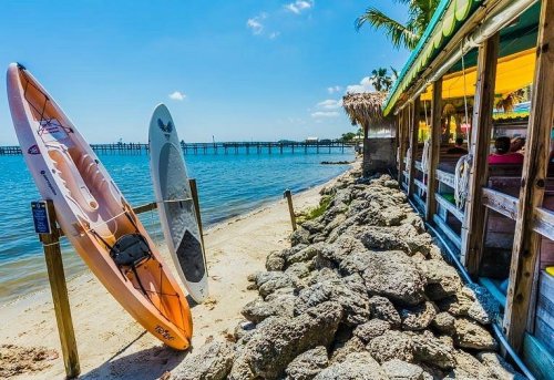 Two kayaks are parked on a sandy beach beside a wooden restaurant, with a pier and clear blue water in the background.