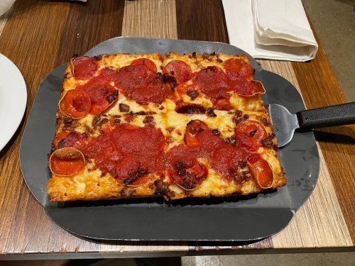 A square pizza topped with pepperoni and cheese, served on a black tray with a pizza cutter beside it.