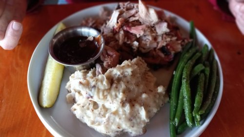 A plate with pulled pork, barbecue sauce, mashed potatoes, green beans, and a pickle slice on a wooden table.