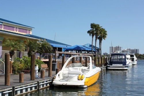 A yellow boat docked by a waterfront restaurant with palm trees and blue umbrellas under a clear sky.
