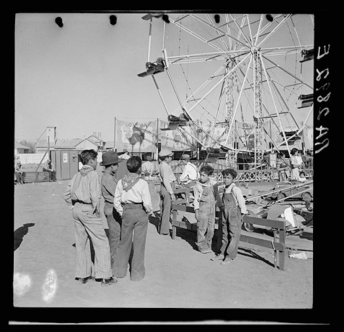A group of children in overalls stands near a Ferris wheel at a fair, with colorful banners in the background.