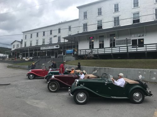Three vintage cars parked in front of a white building with a restaurant sign, people gathered nearby.