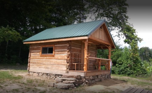 A small wooden cabin with a green metal roof, surrounded by trees and a stone foundation.