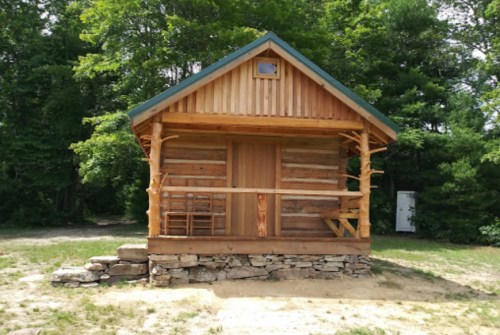 A small wooden cabin with a porch, surrounded by trees and greenery.