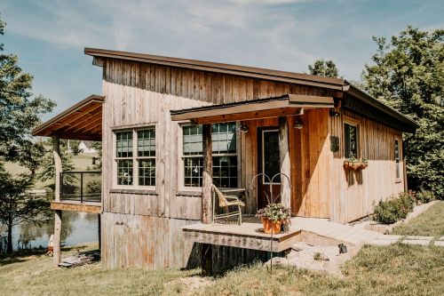 A rustic wooden cabin with large windows, a porch, and a scenic view of greenery and water nearby.