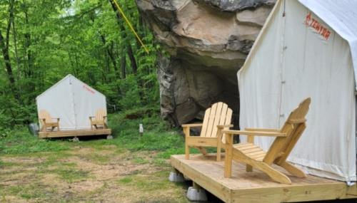 Two white canvas tents on wooden platforms, with Adirondack chairs in front, set in a green forest near a large rock.