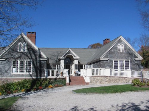 A large, stylish house with a gray shingle exterior, white railings, and a landscaped yard under a clear blue sky.