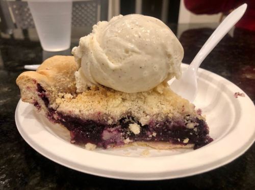 A slice of berry pie topped with a scoop of vanilla ice cream on a paper plate.