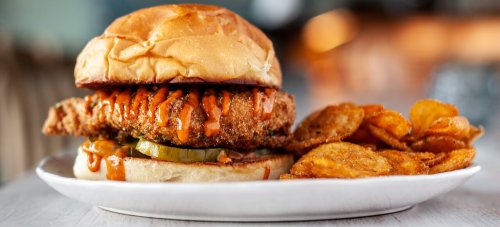 A crispy chicken sandwich with sauce and pickles, served with a side of crispy potato chips on a white plate.