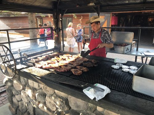 A chef in a hat grills various meats on a large barbecue, with people socializing in the background.