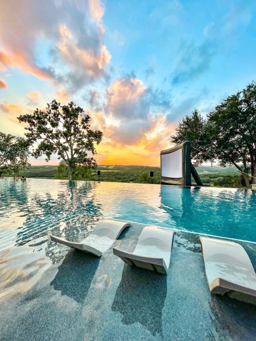 A serene infinity pool at sunset, with lounge chairs and a large screen set against a scenic landscape.