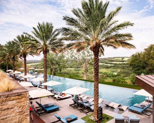 A serene pool area with palm trees, lounge chairs, and umbrellas overlooking lush green hills under a blue sky.