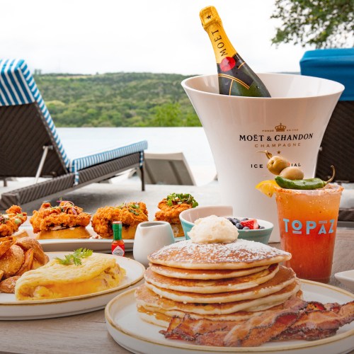 A luxurious brunch spread featuring pancakes, eggs, fried chicken, a cocktail, and champagne by a scenic poolside.