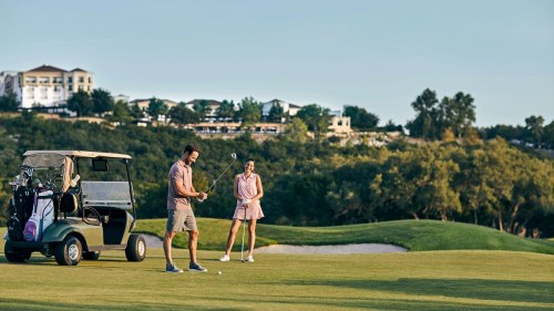 A man prepares to swing a golf club while a woman watches, with a golf cart nearby and a scenic background.