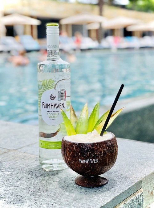A coconut drink with a straw and pineapple leaves sits beside a bottle of RumHaven, by a pool with lounge chairs.