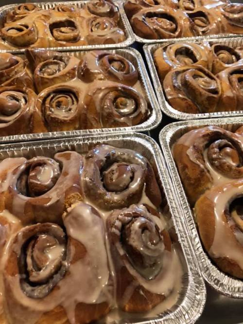 Trays of freshly baked cinnamon rolls drizzled with creamy icing, arranged in aluminum pans.