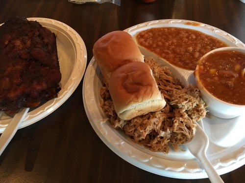 A plate with smoked ribs, pulled pork, two buns, baked beans, and a bowl of chili on a wooden table.