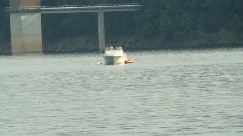 A boat glides across a calm lake, with a bridge and trees visible in the background.