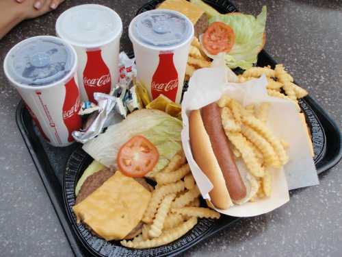 A tray with burgers, a hot dog, crinkle fries, and three cups of Coca-Cola.