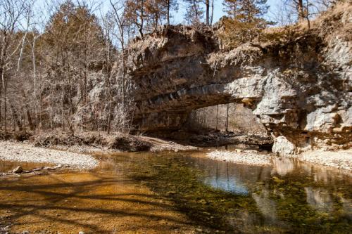 A natural stone arch over a clear stream, surrounded by bare trees and rocky terrain in a serene landscape.