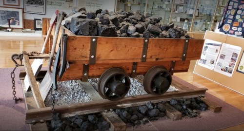 A wooden coal cart filled with rocks, displayed on a gravel track in a museum setting.