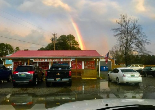 A coffee shop with a red roof under a rainbow, surrounded by parked cars and a cloudy sky.