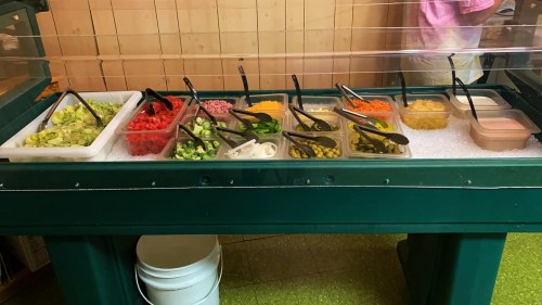 A salad bar with various toppings in containers, including lettuce, tomatoes, olives, and dressings on ice.