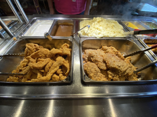 A buffet display featuring fried chicken, mashed potatoes, gravy, and various sides in metal trays.