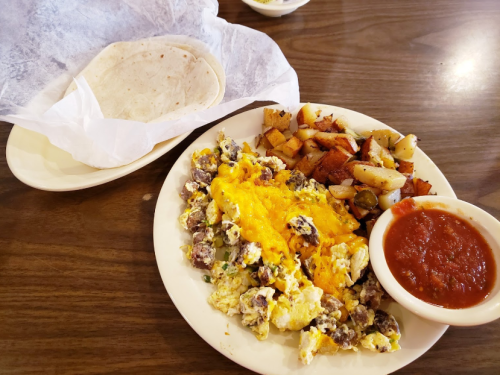 A plate of scrambled eggs with cheese and sausage, seasoned potatoes, a side of salsa, and two tortillas on a table.