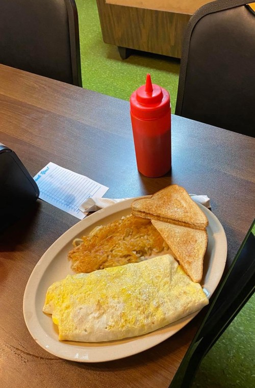 A plate with an omelet, hash browns, and toast, accompanied by a bottle of ketchup on a wooden table.