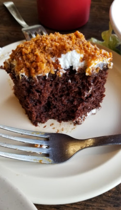 A slice of chocolate cake topped with whipped cream and a crumbly topping, served on a white plate with a fork.