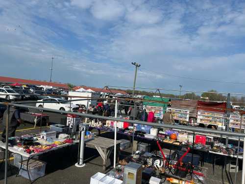 A bustling outdoor market with various stalls selling goods, surrounded by cars and a cloudy sky.