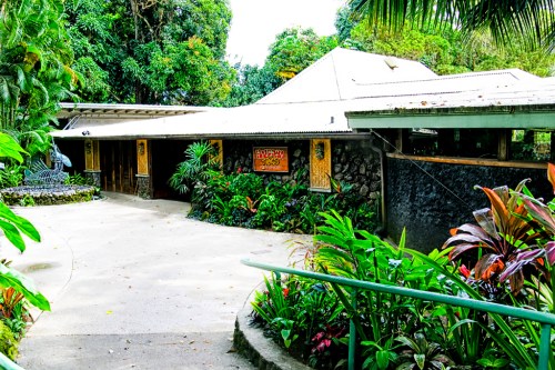 A tropical building surrounded by lush greenery and colorful plants, featuring a stone wall and a sloped roof.