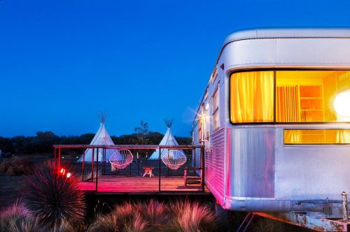 A vintage trailer with warm lights, a deck, and two hanging chairs, set against a twilight sky with teepees in the background.