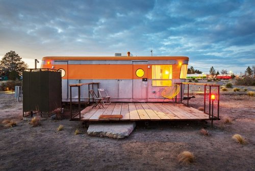 A retro trailer with orange accents sits on a wooden platform in a desert landscape under a cloudy sky.