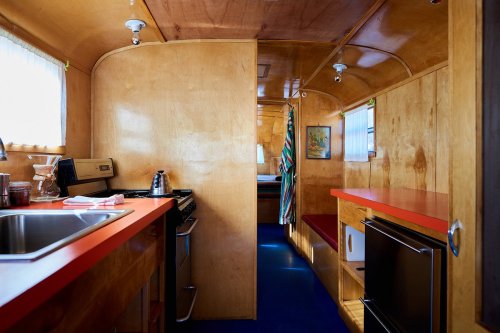 A cozy, vintage kitchen with wooden walls, red countertops, and a view into a small living area.